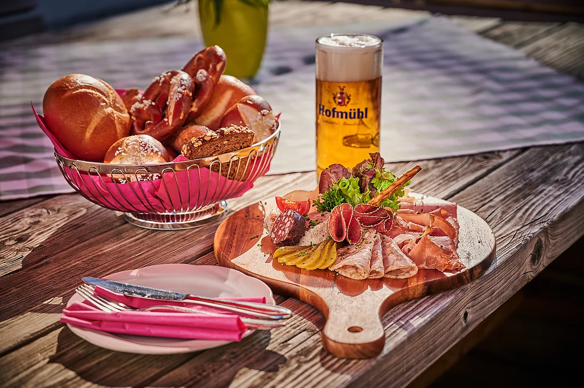Ein gemütliche Brotzeit im Biergarten.
Hierzu wird ein Brotzeitbrettl mit einem Korb Brezen/Semmeln und einem Bier serviert.