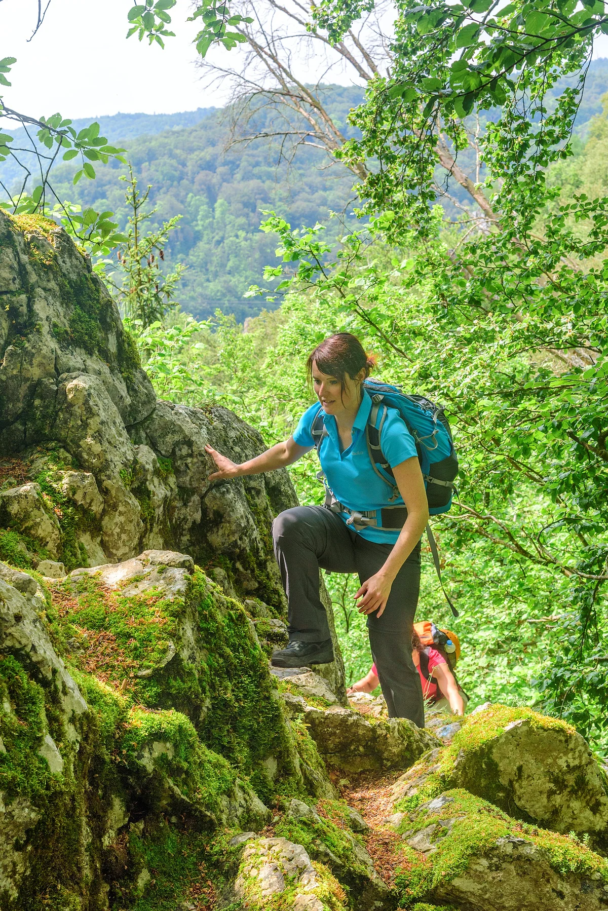 Eine Wanderin mit blauem Shirt und Rucksack wandert entlang Felsen, die mit Moos bewachsen hinauf. Weiter unten ist eine zweite Frau in rosa zu sehen. Hinter ihnen ein schöner Blick ins Tal umgeben von grünem Blätterwerk.