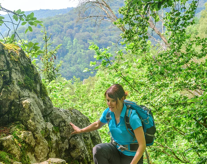 Eine Wanderin mit blauem Shirt und Rucksack wandert entlang Felsen, die mit Moos bewachsen hinauf. Weiter unten ist eine zweite Frau in rosa zu sehen. Hinter ihnen ein schöner Blick ins Tal umgeben von grünem Blätterwerk.