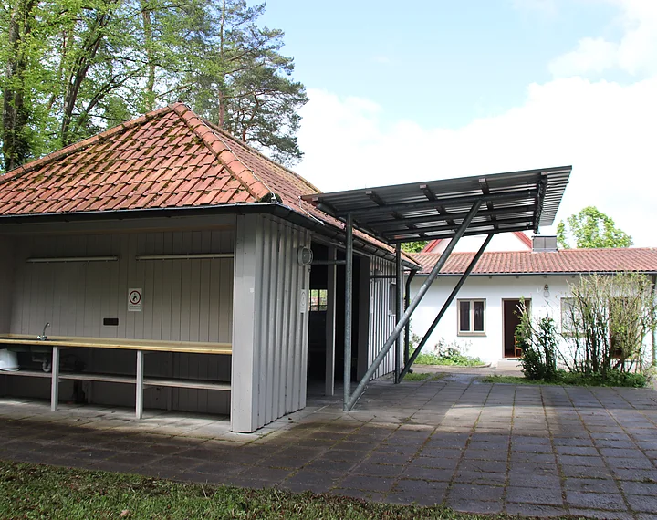 Ein kleiner, überdachter Waschplatz mit Spüle und Arbeitsfläche im Freien, umgeben von Bäumen.