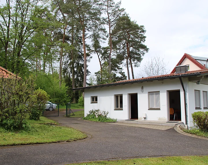 Ein einstöckiges weißes Gebäude mit mehreren Fenstern und offenen Türen, umgeben von Bäumen und einem gepflasterten Weg.