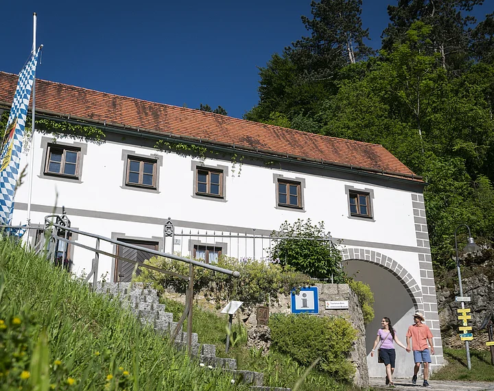 Weißes Gebäude mit rotem Ziegeldach, bayerischer Flagge und zwei Personen vor einem Torbogen bei Sonnenschein