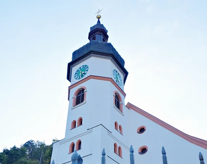 Wellheim Kath. Pfarrei St. Andreas Außenansicht Blick durch ein Metalltor auf eine weiße Kirche mit Turmuhr und Treppe, im Hintergrund Bäume.