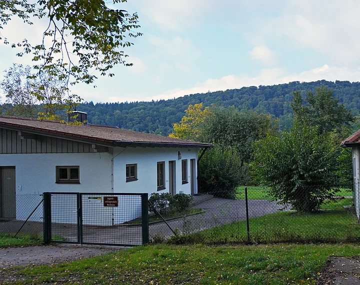 Eingezäuntes Grundstück mit zwei Gebäuden, Bäumen und bewaldetem Hügel im Hintergrund bei Tageslicht.